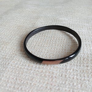 May include: A black bangle bracelet with a copper-coloured accent. The bracelet is circular and appears to be made of wood or a similar material. The copper accent adds a touch of contrast to the otherwise dark design.