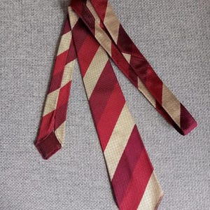 May include: A necktie featuring a diagonal striped pattern in alternating shades of deep burgundy, bright red, and light beige. The fabric has a visible woven texture, and the tie is folded to display its pointed end and the length of the blade.