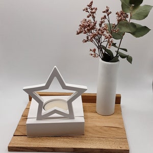 Op de afbeelding: Witte houten stervormige kaarsenhouder met een kleine witte kaars erin, die op een houten dienblad staat met een witte keramische vaas met roze bloemen.