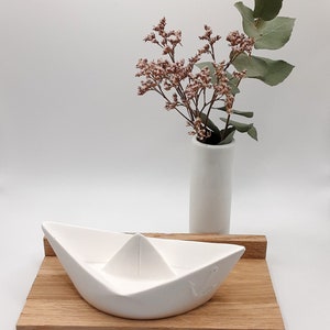 May include: A white ceramic paper boat with an anchor design sits on a wooden tray. A white vase with pink and green flowers is behind the boat.