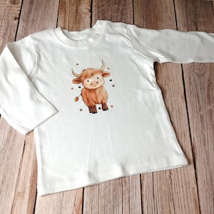 Könnte beinhalten: Weißes Langarmshirt mit einer braunen und weißen Comic-Highland-Kuh mit schwarzen Augen und einer braunen Nase. Die Kuh ist von kleinen braunen Punkten umgeben.
