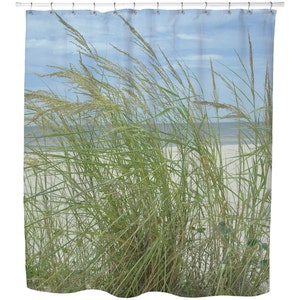 May include: Shower curtain featuring a beach scene with tall green and tan grasses blowing in the wind. The background shows a sandy beach, ocean, and a blue sky with clouds. The curtain is hung on a white rod.