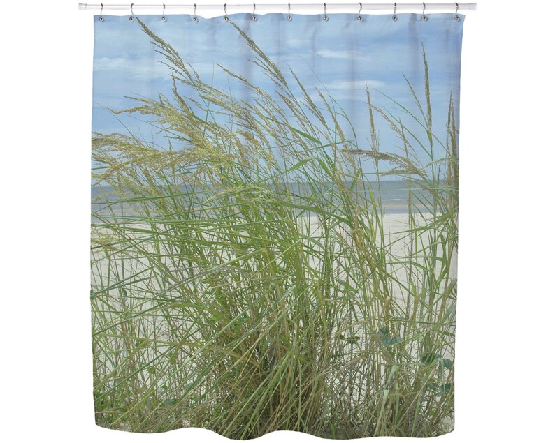 May include: Shower curtain featuring a beach scene with tall green and tan grasses blowing in the wind. The background shows a sandy beach, ocean, and a blue sky with clouds. The curtain is hung on a white rod.