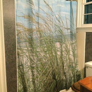May include: A shower curtain featuring a coastal scene with tall green and tan grasses blowing in the wind against a blue sky. The curtain is hanging in a bathroom with a window and a toilet.