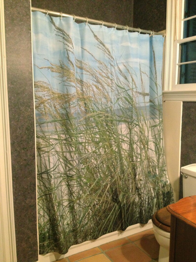 May include: A shower curtain featuring a coastal scene with tall green and tan grasses blowing in the wind against a blue sky. The curtain is hanging in a bathroom with a window and a toilet.