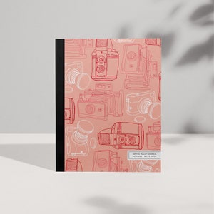 Può includere: Un diario a punti rosa con una rilegatura nera. La copertina presenta un motivo ripetuto di disegni di macchine fotografiche vintage in rosso.