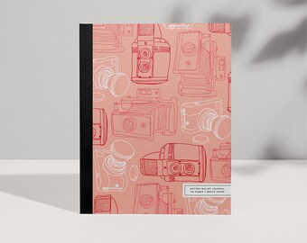 Puede incluir: Un diario de puntos rosa con una encuadernación negra. La portada presenta un patrón repetido de dibujos de cámaras vintage en rojo.