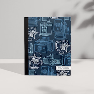 Può includere: Un diario a punti blu con un contorno bianco di macchine fotografiche vintage sulla copertina. Il testo sulla copertina recita "Dotted Bullet Journal 110 Pages | White Paper".
