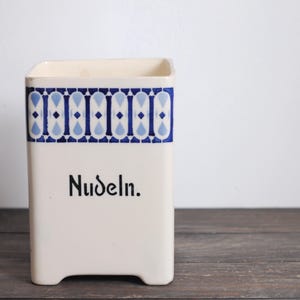 May include: A rectangular, off-white ceramic container with a blue and white geometric pattern around the top. The word "Nudeln" is printed in black on the front. The container has a slightly flared opening and a small base.