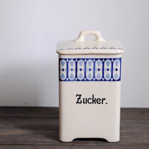 May include: Cream-colored ceramic sugar container with a rectangular shape and a matching lid. The container features a blue and white geometric pattern around the top and the word "Zucker" in black script.