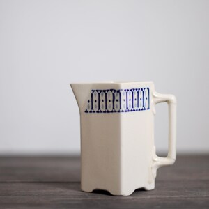 May include: Cream-colored ceramic pitcher with a geometric design in blue. The pitcher has a square shape with a handle and a spout. The blue design is a repeating pattern of dots and lines around the top of the pitcher.