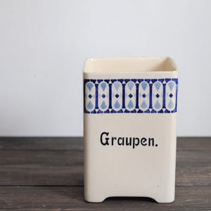 May include: A rectangular, off-white ceramic container with a blue and white geometric pattern around the top. The word "Graupen" is printed in black script on the front. The container has a slightly curved top edge and sits on a dark wooden surface.