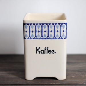 May include: Cream-colored ceramic container with a blue and white geometric pattern around the top. The word "Kaffee" is printed in black on the front. The container has a rectangular shape with a slightly flared top.