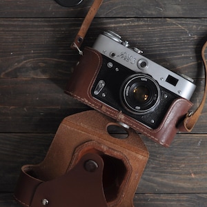 May include: A classic silver and black camera with a brown leather case and strap. The camera has a lens and is labelled "FED 3". The case is open, revealing the camera. A black lens cap is also present.