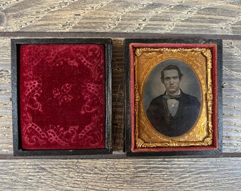 May include: A vintage photograph of a man in a dark suit and bow tie, framed in a gold-colored frame with a red velvet mat. The photograph is in a wooden case with a red velvet lining.