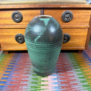 Puede incluir: Un jarrón de cerámica hecho a mano con un esmalte moteado verde oscuro y negro. El jarrón tiene forma cilíndrica con ranuras horizontales y un borde ligeramente ensanchado. El jarrón mide unos 18 cm de alto.