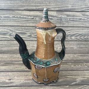 Puede incluir: Una tetera de cerámica marrón y negra con un esmalte único y moteado. La tetera tiene una tapa alta y puntiaguda y un pico curvo.