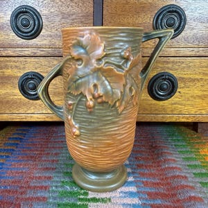 May include: A ceramic pitcher featuring a textured, horizontal ribbed surface in shades of brown and olive green. The vessel is adorned with a raised relief of grape leaves and clusters of grapes, and includes two angular handles on opposite sides.