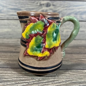 May include: A small brown ceramic pitcher with a green handle. The pitcher has a design of colorful leaves on the front.