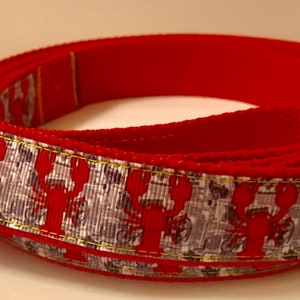 Peut inclure: Une laisse rouge pour chien avec un motif répété de homards rouges sur un fond gris et blanc. La laisse a un dos rouge uni et une quincaillerie en métal argenté.