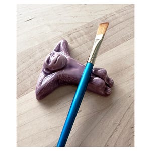 May include: A purple ceramic monkey figurine with a relaxed pose, holding a blue and silver paintbrush. The monkey is a brush holder.