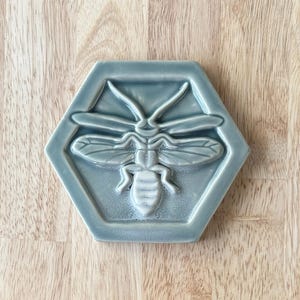 May include: A hexagonal ceramic tile with a raised, white bee design in the center. The tile is a light blue-gray color.