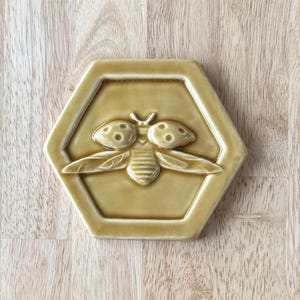 May include: A ceramic tile with a raised bee design inside a hexagonal frame. The bee is detailed with wings, antennae, and body segments. The tile is a muted yellow-brown color, and the background is a light wood grain.