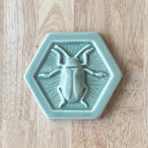 May include: A light blue ceramic tile with a raised design of a beetle. The beetle is in the center of the tile and is surrounded by a leafy design.