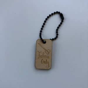 May include: A wooden tag with the text "Fabric Only" and a scissors graphic, attached to a black beaded chain. The tag is rectangular with rounded corners and a light brown color.