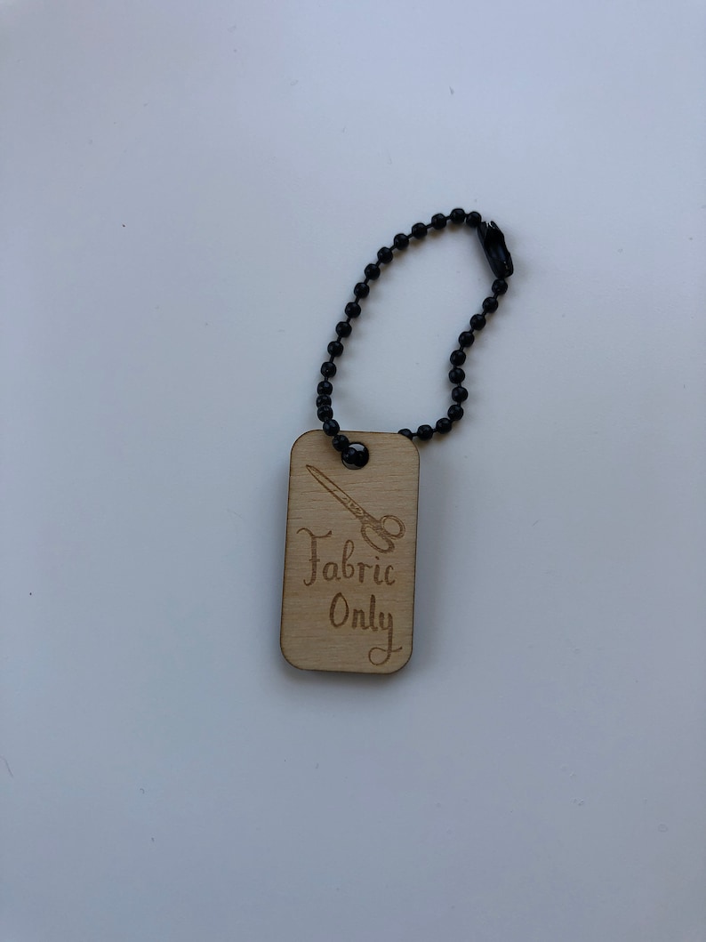 May include: A wooden tag with the text "Fabric Only" and a scissors graphic, attached to a black beaded chain. The tag is rectangular with rounded corners and a light brown color.