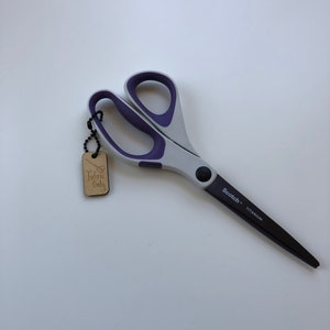 May include: Gray and purple fabric scissors with black titanium blades. The scissors have a gray handle with purple accents and a black pivot screw. A wooden tag that says "Fabric Only" is attached to the scissors.