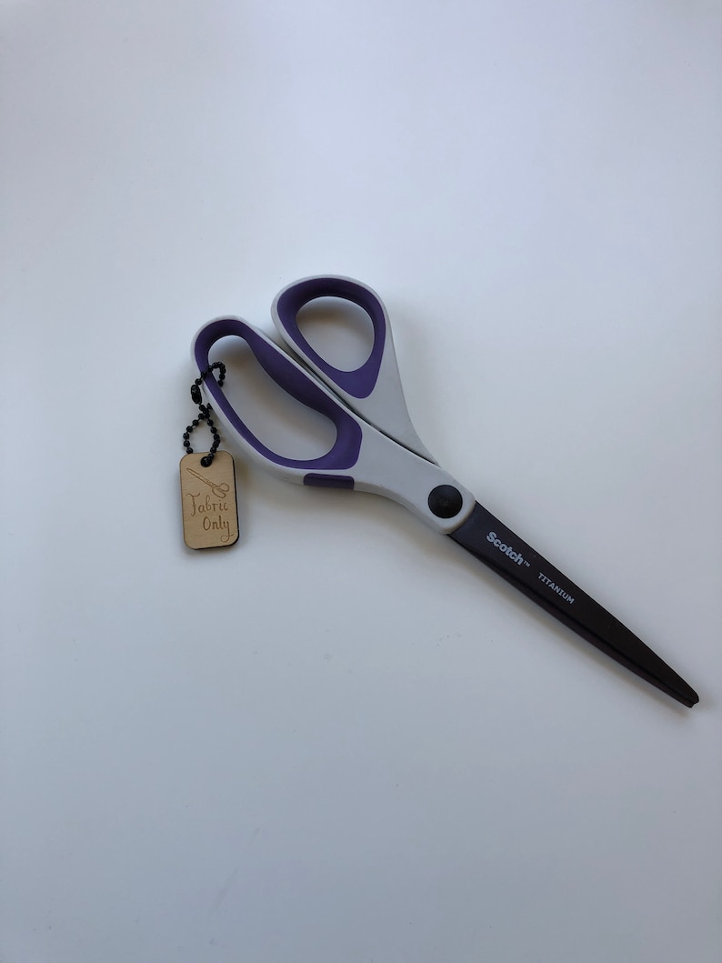 May include: Gray and purple fabric scissors with black titanium blades. The scissors have a gray handle with purple accents and a black pivot screw. A wooden tag that says "Fabric Only" is attached to the scissors.