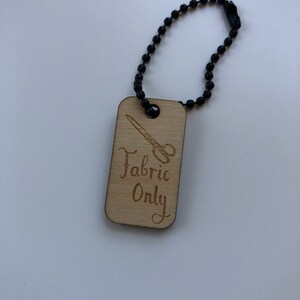 May include: A rectangular wooden tag with the words "Fabric Only" and a scissors graphic, attached to a black ball chain. The text and graphic are etched into the light-colored wood.
