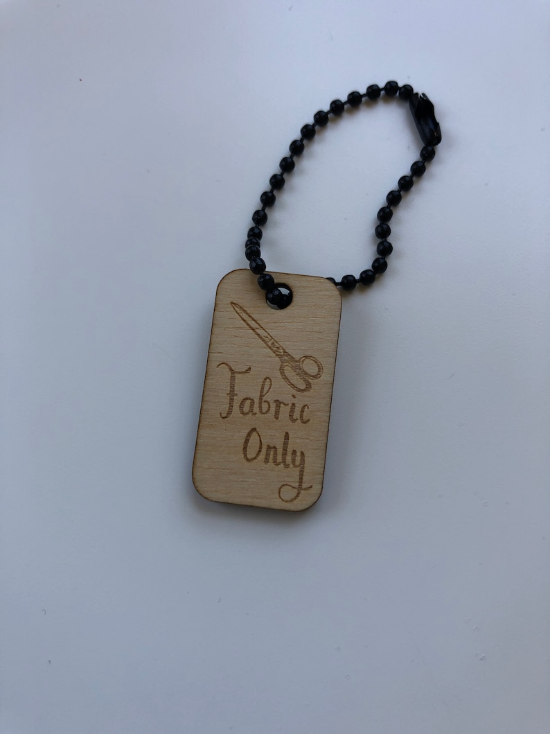 May include: A rectangular wooden tag with the words "Fabric Only" and a scissors graphic, attached to a black ball chain. The text and graphic are etched into the light-colored wood.