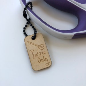 May include: A wooden tag with the words "Fabric Only" and a scissors graphic, attached to a pair of purple and white scissors with a black beaded chain. The tag is light brown, and the scissors have a curved handle.