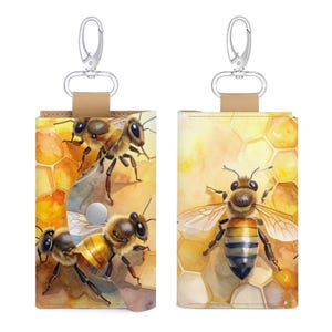 May include: A rectangular leather key case featuring a watercolour illustration of honeybees on a honeycomb background. The case has a silver metal snap button closure and a silver swivel lobster clasp attached to a tan leather strap at the top.