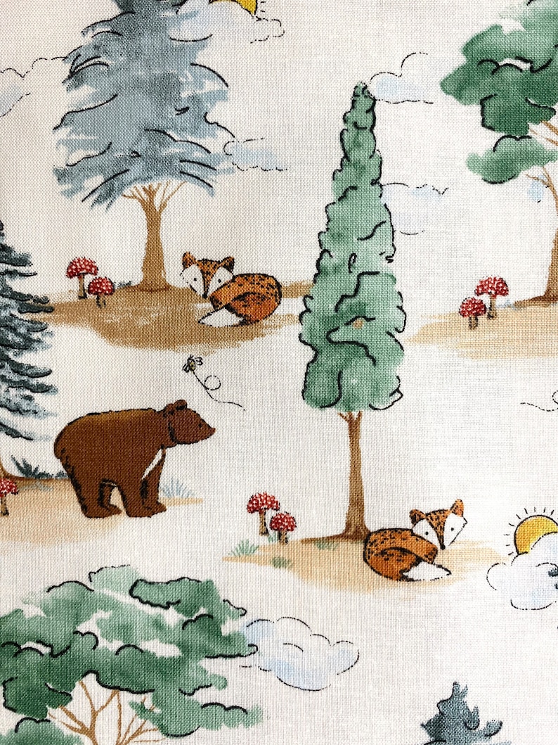 May include: A white fabric with a repeating pattern of green trees, brown bears, orange foxes, red mushrooms, and blue clouds. The background is a light cream color.