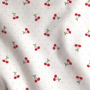 May include: A white fabric featuring a repeating pattern of small red cherries with green stems and leaves, interspersed with tiny black polka dots. The material has a soft, folded texture that creates subtle shadows across the surface.