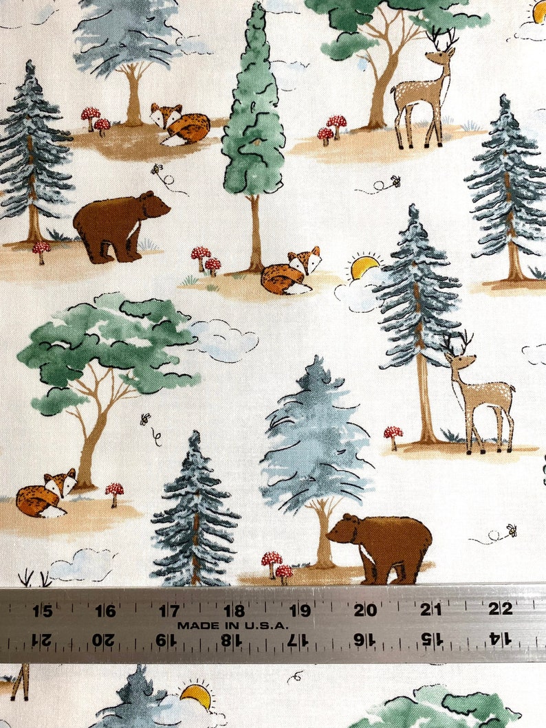 May include: White fabric with a repeating pattern of brown bears, foxes, deer, and pine trees. The background is a light beige with brown and green accents. The fabric is measured with a ruler in inches.