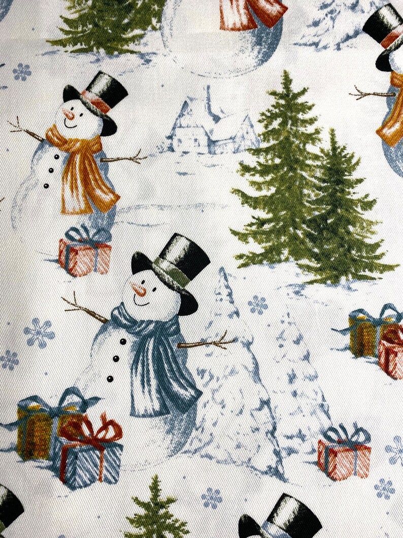Puede incluir: Tela blanca con un patr&oacute;n repetido de mu&ntilde;ecos de nieve, pinos, copos de nieve y regalos envueltos. Los mu&ntilde;ecos de nieve llevan sombreros de copa negros y bufandas en tonos naranja y azul. Los regalos est&aacute;n envueltos en rojo, azul y dorado.