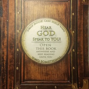 May include: A vintage-style book cover with a wooden background and gold accents. The cover features a circular design with the text "THIS BOOK CAN HELP YOU HEAR GOD SPEAK TO YOU! OPEN THIS BOOK ANYWHERE AND KEEP READING UNTIL YOU FIND SOMETHING..."