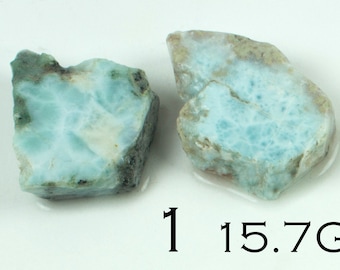 May include: Two pieces of raw larimar gemstone, displaying a light blue color with white and brown inclusions. The stones have a rough, natural texture. The text "1 15.7G" is visible.