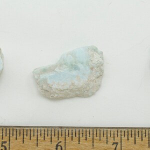 May include: Three rough-cut larimar stones with a light blue and white color pattern. The stones are irregularly shaped and vary in size. The background is white, and a ruler is at the bottom of the image.