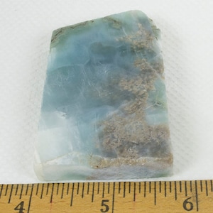 May include: A rectangular piece of polished larimar gemstone. The stone displays a blend of light blue and white hues, with areas of brown and tan veining. A wooden ruler with inch markings is visible along the bottom edge, with measurements from 4 to 6 inches.