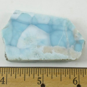 May include: A piece of polished larimar gemstone, showcasing a blend of light blue and white hues with a marbled appearance. The stone has a natural, irregular shape and is set against a white background. A wooden ruler is placed below the stone.