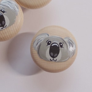 Könnte beinhalten: Vier Holzknöpfe mit bemalten Koala-Gesichtern. Die Koalas haben graues Fell, schwarze Nasen und schwarze Augen. Die Knöpfe sind hellbraun.