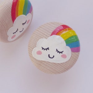 Könnte beinhalten: Zwei Holzknöpfe, die mit einem Regenbogen und einer weißen Wolke mit einem lächelnden Gesicht bemalt sind. Die Wolke hat geschlossene Augen und rosige Wangen.