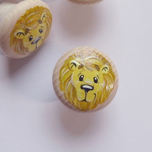 Könnte beinhalten: Vier Holzknöpfe mit bemalten Löwenköpfen. Die Löwen haben gelbes Fell, schwarze Augen und rosa Nasen. Die Knöpfe sind hellbraun.