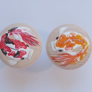 Könnte beinhalten: Zwei runde Holzknöpfe mit bemalten Koi-Fisch-Designs. Ein Knopf zeigt einen roten und schwarzen Koi-Fisch, während der andere Knopf einen orangefarbenen und weißen Koi-Fisch zeigt.
