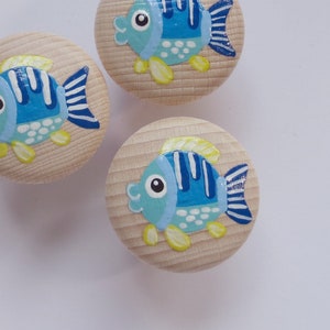 Könnte beinhalten: Drei Holzknöpfe mit bemalten blauen und gelben Fischmotiven. Die Fische haben weiße Punkte und blaue Streifen. Die Knöpfe sind rund und haben eine helle Holzoptik.